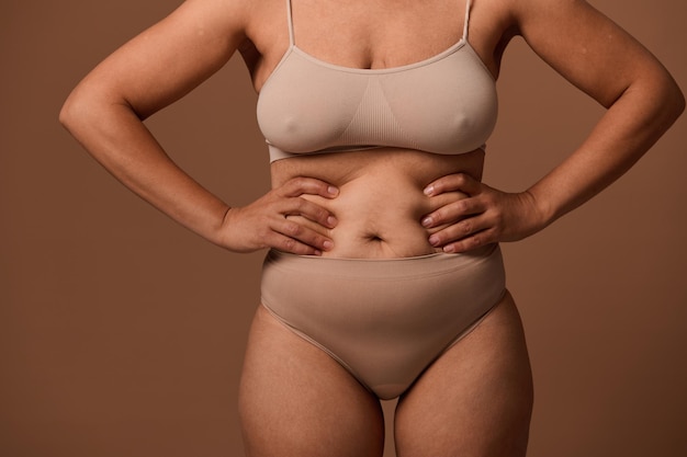 Vista ritagliata di una donna irriconoscibile in biancheria intima beige che posa le mani sullo stomaco con muscoli rilassati che hanno perso il tono e le smagliature dopo la gravidanza e il parto