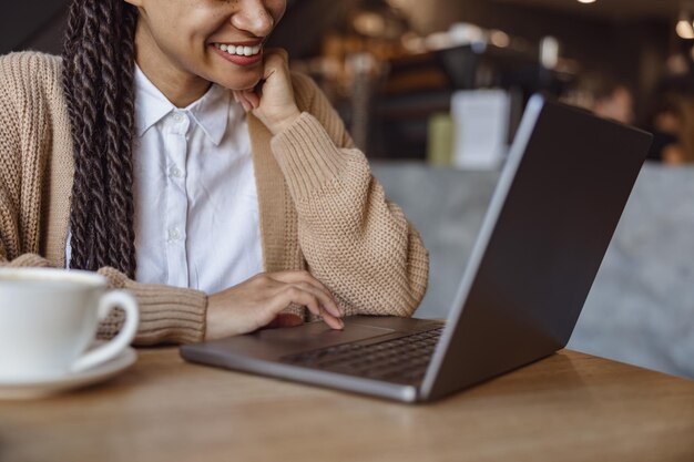 Vista ritagliata su una donna etnica carina che sorride sorriso a trentadue denti mentre lavora al computer portatile durante la pausa caffè all'interno di una caffetteria