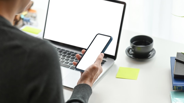 Vista sopra la spalla del giovane libero professionista che utilizza il telefono cellulare e lavora con il computer portatile sul posto di lavoro moderno.