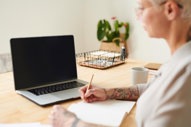 Vista sopra la spalla di una donna moderna con tatuaggi seduta alla scrivania e facendo appunti nel diario mentre studia marketing online