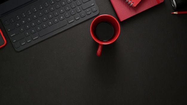 Vista superiore del posto di lavoro alla moda con tastiera wireless, articoli per ufficio, copia spazio e tazza di caffè rosso sul tavolo nero