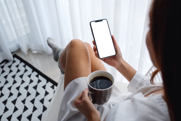 Vista superiore del telefono cellulare della tenuta della donna con lo schermo da tavolino bianco in bianco mentre sedendosi e bevendo caffè in camera da letto