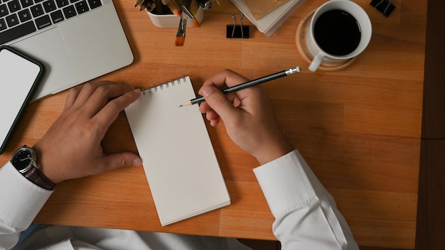 Vista superiore della mano maschio che scrive sul taccuino in bianco sul tavolo da lavoro in legno in ufficio a casa