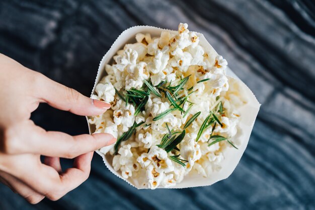 Vista superiore della mano pizzicando un popcorn di rosmarino in sacchetto di carta sul tavolo di marmo superiore.