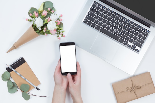 Vista superiore delle mani della donna con il cellulare con lo schermo bianco, computer portatile, telefono cellulare, composizione nel fiore. Disposizione piana dell'area di lavoro