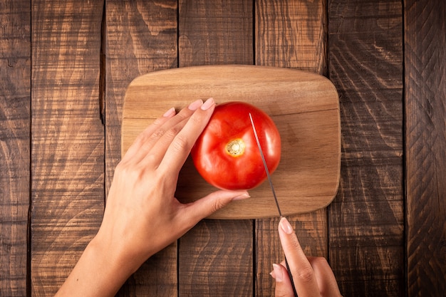 Vista superiore delle mani di una donna che tagliano con un coltello un pomodoro su una tavola su un tavolo di legno