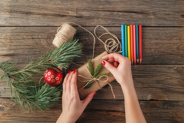 Vista superiore delle mani femminili che tengono regalo o presente di natale. Regali e pergamene confezionati, rami di abete e strumenti sul tavolo di legno squallido. Luogo di lavoro per la preparazione di decorazioni fatte a mano.