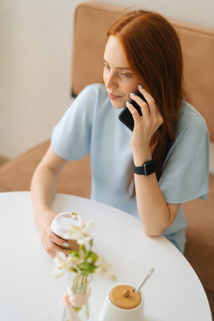 Vista verticale dall'alto di una giovane donna felice che tiene in mano una tazza con caffè caldo in mano che parla al telefono cellulare seduto alla scrivania del bar. Signora caucasica della testarossa graziosa che ha attività per il tempo libero nella caffetteria.