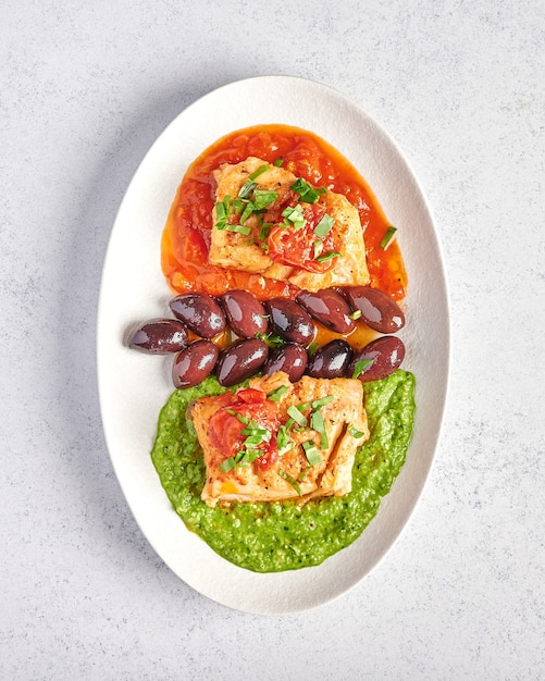 Vista verticale pesce bianco con olive, pesto e salsa di pomodori e peperoni al forno sulla piastra ovale bianca, concetto di stile alimentare, vicino, spazio di copia