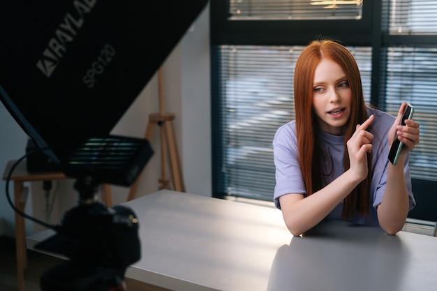 Vista webcam di una giovane blogger sorridente che saluta i follower e parla del telefono durante la chat video sullo sfondo della finestra. POV di donna rossa allegra che ha presentazione durante il tramonto.