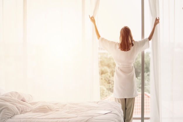 Visualizza indietro le tende di apertura della donna in una camera da letto bianca