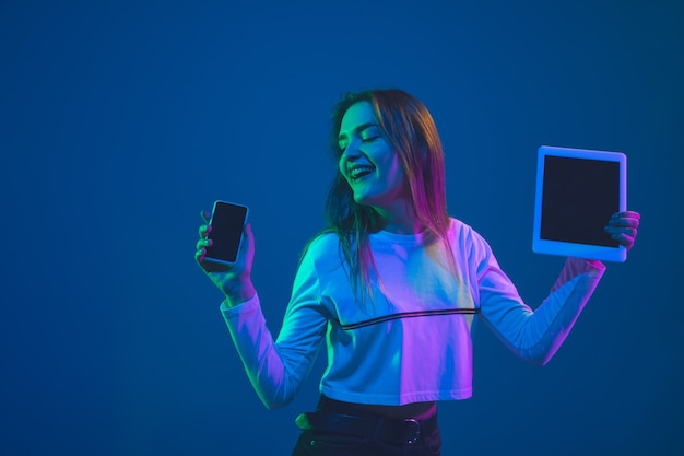 Visualizzazione dei dispositivi. Ritratto di giovane donna caucasica isolato su sfondo blu studio in luce al neon. Bellissimo modello femminile. Concetto di emozioni umane, espressione facciale, vendite, pubblicità, cultura giovanile.