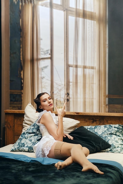 Vita boema Attrice in pigiama vintage in camera da letto sdraiata su un letto con un bicchiere di vino rosso