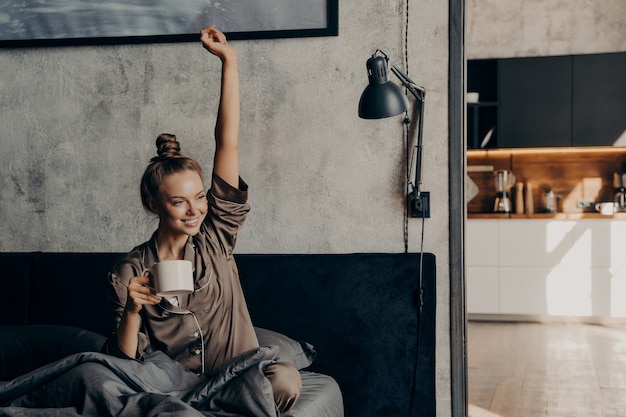 Vita felice. Attraente giovane donna felice che ha il caffè mattutino a letto dopo essersi svegliato, vestita con un pigiama di raso, allungando la mano e godendosi il nuovo giorno mentre si rilassa nel fine settimana a casa