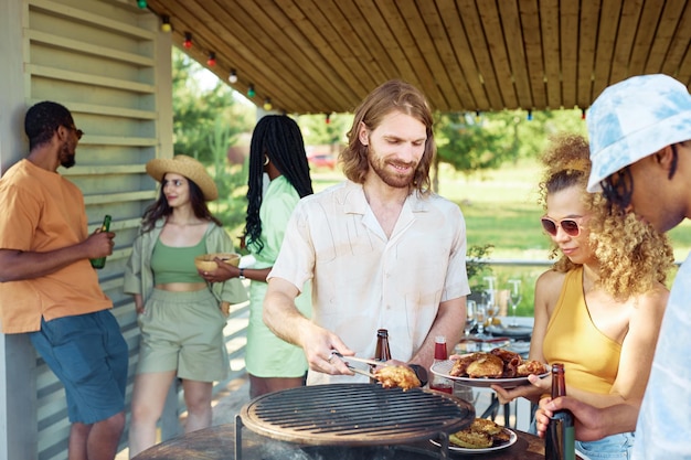 Vita in alto ritratto di uomo sorridente che griglia carne all'aperto durante la festa barbecue estiva con gli amici