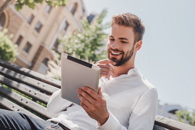 Vita sociale giovane uomo sorridente seduto su una panchina nel parco con in mano il suo tablet