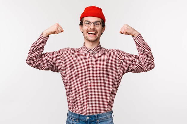 Vita-up ritratto di giovane ragazzo magro fiducioso incoraggiato ed energizzato in berretto rosso e occhiali flettono i bicipiti, mostrando i suoi muscoli forti e potenti