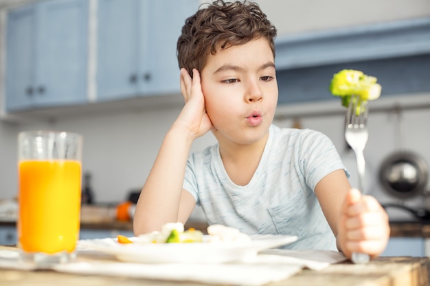 Voglio i biscotti. Ragazzino dai capelli scuri sconvolto bello fare colazione sana e bere un po 'di succo e guardando la verdura verde sulla sua forchetta