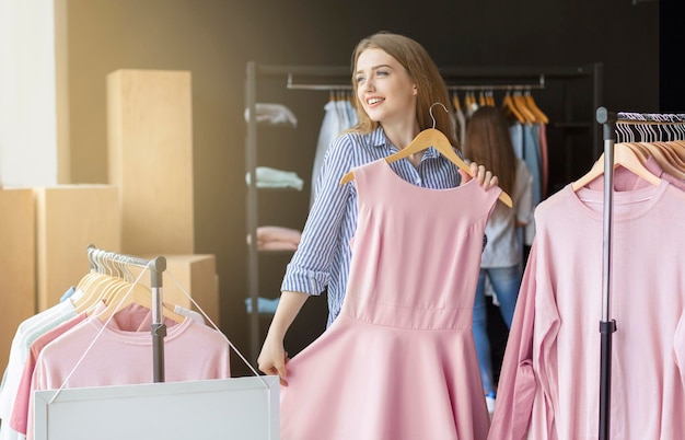 Voglio indossare questo vestito elegante. Ragazza caucasica sognante che prova un vestito rosa al negozio di abbigliamento, copia spazio