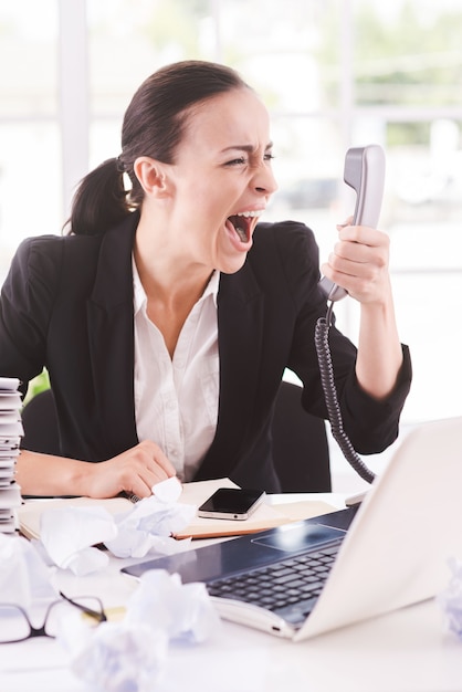 Voglio quel rapporto adesso! Furiosa giovane imprenditrice in abiti da cerimonia che grida al telefono mentre è seduta al suo posto di lavoro