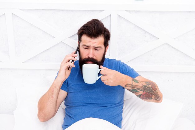 Voglio solo sorseggiare un caffè Uomo barbuto parlare al telefono bere caffè a letto Bere abitudine al tè Bere cacao Divertirsi a bere tè o altre bevande calde Vita moderna Routine mattutina