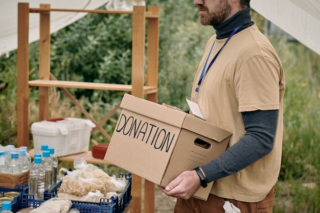 Volontario barbuto con distintivo che trasporta scatola di cartone con donazione a tavola con merce per senzatetto