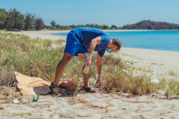 Volontario in maschera facciale blu lazur mare spiaggia di sabbia uomo attivo raccogliere immondizia plastica sacchetti di schiuma film spazzatura versata spazzatura pianeta inquinamento protezione ambientale problemi sociali naturali concetto