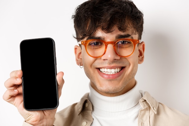 Volto di bel ragazzo sorridente in vetri che mostra ghiaione smartphone vuoto, muro bianco.