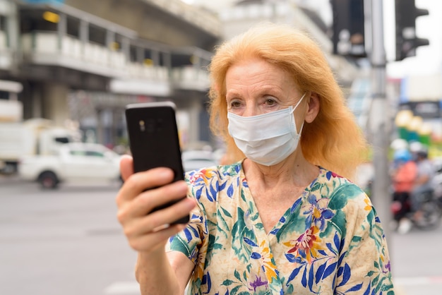 Volto di donna bionda senior con maschera utilizzando il telefono in città all'aperto