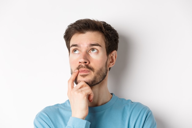 Volto di giovane uomo caucasico alzando lo sguardo pensieroso, facendo una scelta o pensando, meditando stando in piedi su sfondo bianco.