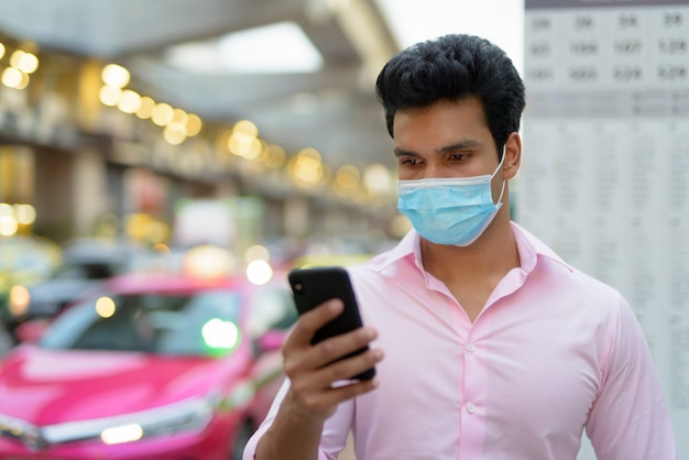 Volto di giovane uomo d'affari indiano con maschera utilizzando il telefono alla fermata dell'autobus