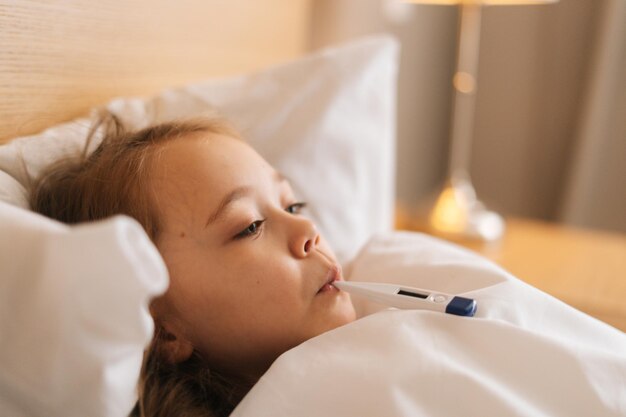 Volto ravvicinato di una bambina stanca che si addormenta in casa sotto una coperta, occhi che sbattono lentamente le palpebre, coperta avvolta in testa. Primo piano di piccolo bambino malato sdraiato a letto nella stanza dei bambini.