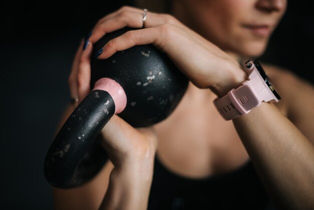 Volto ravvicinato di una bella giovane donna atleta che tiene kettlebell nero con entrambe le mani durante l'allenamento di allenamento sportivo presso la moderna palestra fitness con interni scuri.