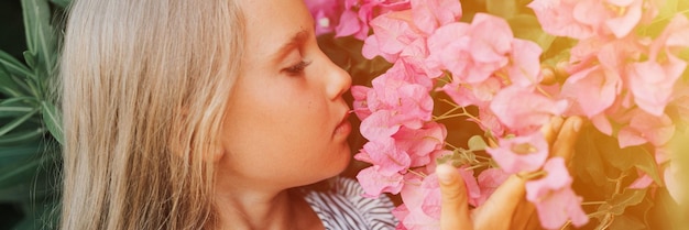 Volto ritratto di candida bambina di otto anni su sfondo di piante verdi e ammirato godersi l'odore dei fiori rosa durante le vacanze estive viaggio gen z concetto di salute mentale banner bagliore