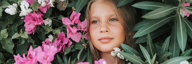 Volto ritratto di candida bella bambina di otto anni con occhi marroni su sfondo di piante verdi e fiori rosa durante una vacanza estiva viaggio gen z concetto di salute mentale banner