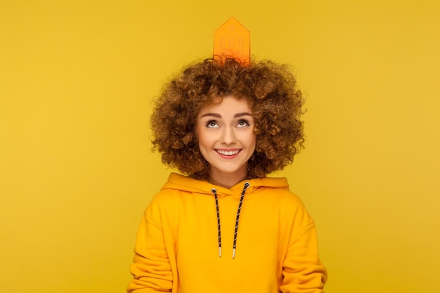 Vorrei casa. Ritratto di felice gioiosa giovane donna dai capelli ricci in felpa con cappuccio in stile urbano che guarda la casa di carta sulla testa, sognando il proprio immobile. tiro in studio indoor isolato su sfondo giallo