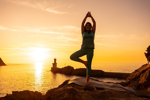 Vrksasana una donna che fa esercizi di meditazione e yoga su una roccia al tramonto accanto a un faro nel mare vita sana e naturista