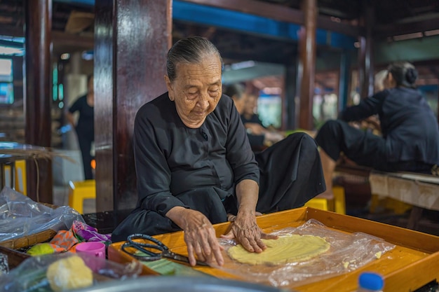 Vung Tau VIETNAM 12 GENNAIO 2023 Focus vecchia donna con abito tradizionale vietnamita ao ba ba Fare avvolgimento Tet Cake il nuovo anno lunare vietnamita Tet cibo all'aperto a mano