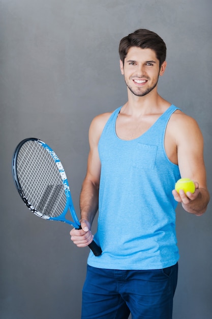 Vuoi giocare con me? Sorridente giovane uomo muscoloso che tiene la racchetta da tennis e allunga la mano con la palla mentre sta in piedi su sfondi grigi