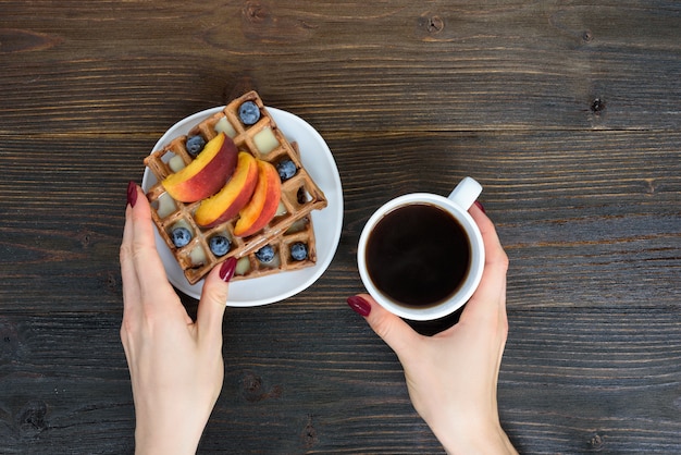 Waffle belgi con frutta e bacche. Mano femminile con una tazza di caffè. Sfondo in legno Vista dall'alto