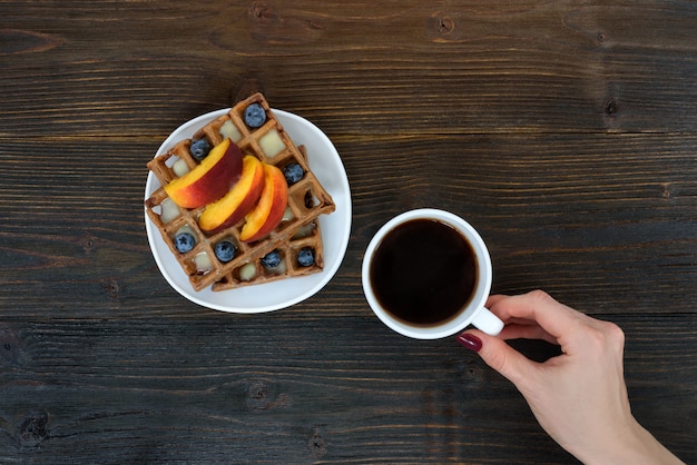 Waffle belgi con frutta e bacche. Mano femminile con una tazza di caffè. Vista dall'alto