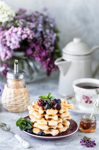 Waffle fatti in casa con frutti di bosco e miele, una tazza di caffè sul tavolo con un mazzo di lillà.