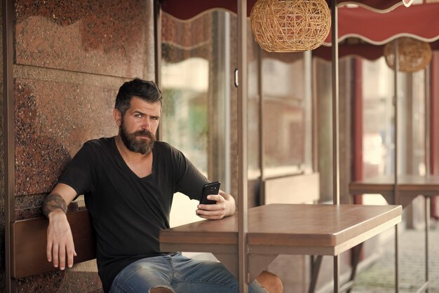 Weblog e sms nuova tecnologia e uomo d'affari con la barba con il telefono cellulare siedono nella caffetteria all'aperto