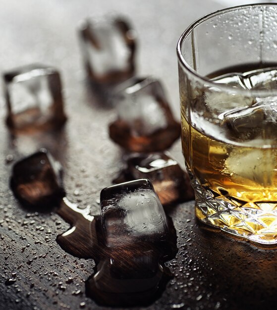 Whisky con ghiaccio con un bicchiere. Cubetti di ghiaccio su un tavolo di legno e un bicchiere con alcol forte refrigerato. Tavolo con whisky americano, bourbon e carte.