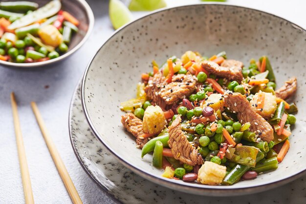 Wok con carne di tacchino, mais, piselli, fagiolini e carote, servito su sfondo grigio con chopstic