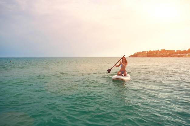 Womanl in bikini blu si siede su una tavola da sup con una pagaia in mare contro il tramonto