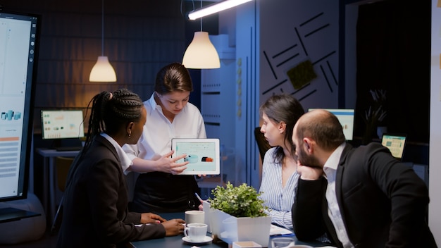 Workaholic focalizzato imprenditrice tenendo tablet che spiega il grafico aziendale superlavoro nella sala riunioni dell'ufficio a tarda notte. Diversi colleghi multietnici che risolvono problemi di statistica in serata