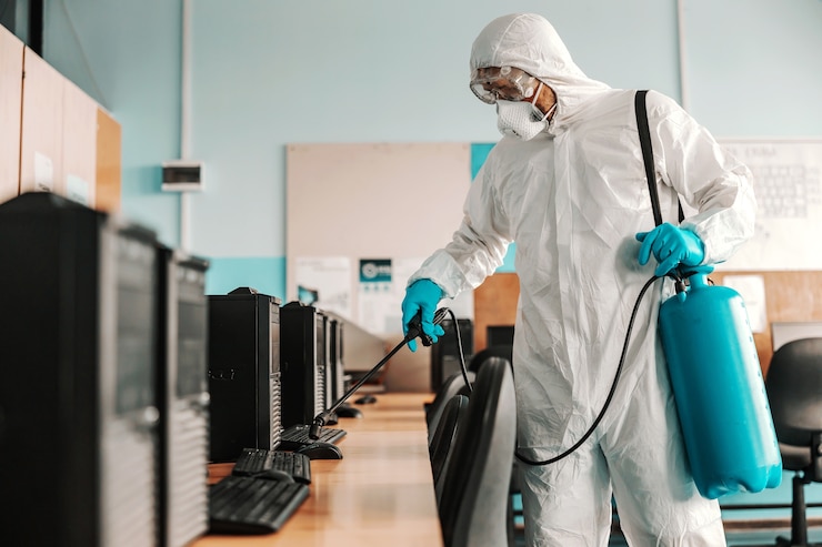 Photo worker in white sterile uniform, with rubber gloves and mask on holding sprayer with disinfectant and sterilizing informatics cabinet in school.