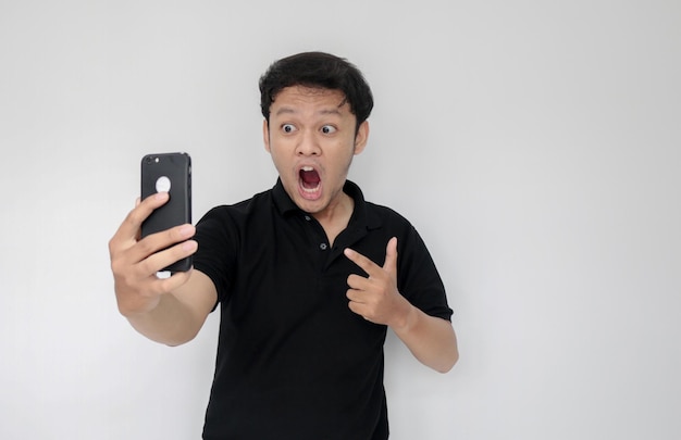 Wow faccia del tuo uomo asiatico ha scioccato ciò che vede nello smartphone su sfondo grigio isolato Indonesia Uomo indossa una camicia nera Sfondo grigio isolato