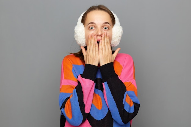 Wow Foto di una giovane donna che indossa paraorecchie bianchi soffici e un maglione colorato sentendosi scioccata e profondamente sorpresa in posa isolata su sfondo grigio che copre la bocca con le palme